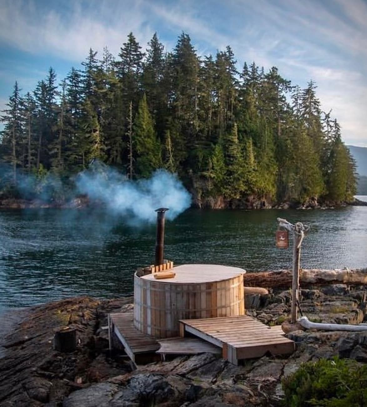 A cedar wood-fired AlumiTubs hot tub steaming on a rocky shoreline, a forested island across the water.