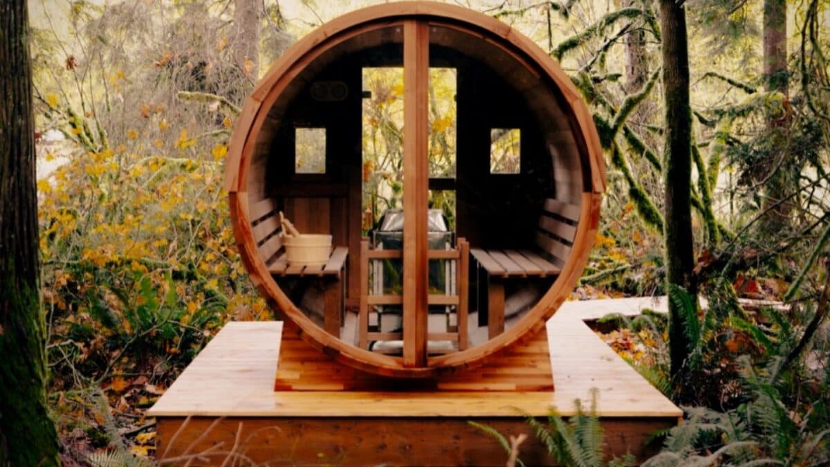 A round cedar barrel sauna with a glass door, centered on a wooden deck in a lush green forest clearing.