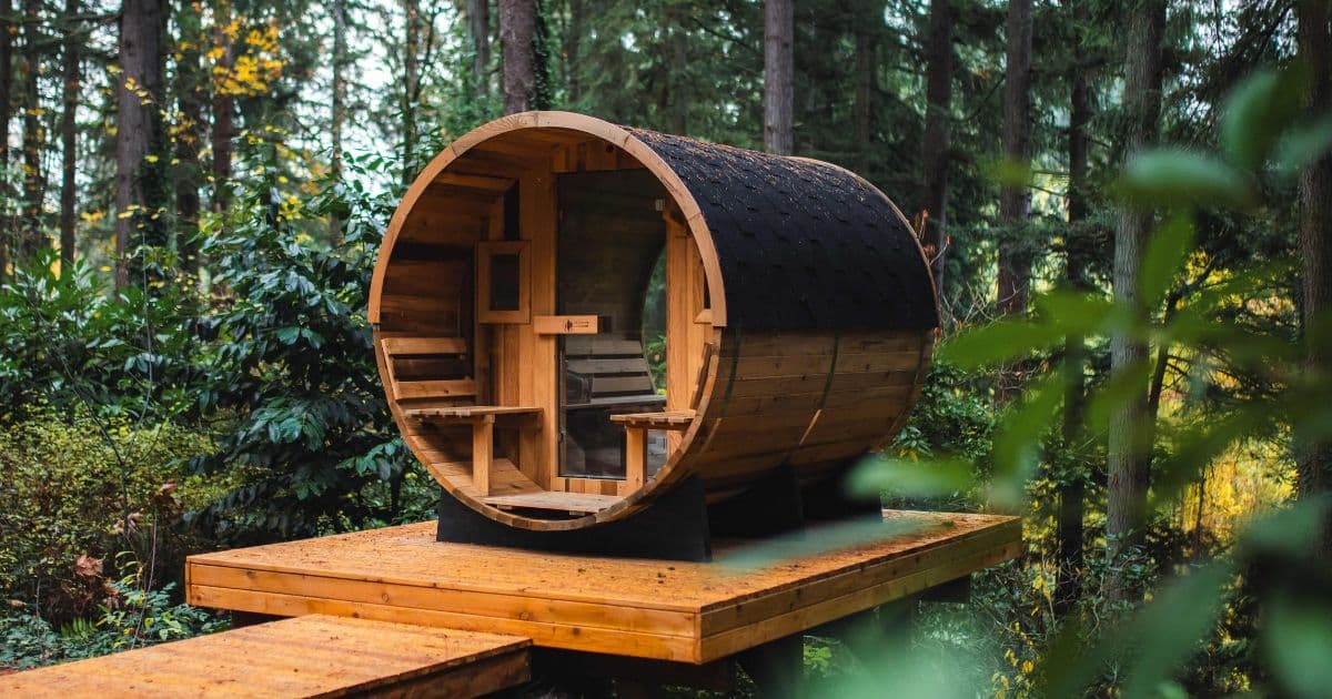 A cedar barrel sauna with a shingled roof on a wooden deck in a lush evergreen forest.