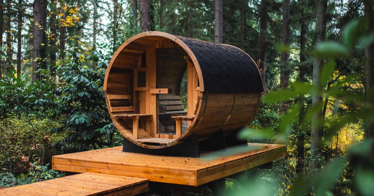 A cedar barrel sauna with a shingled roof on a wooden deck in a lush evergreen forest